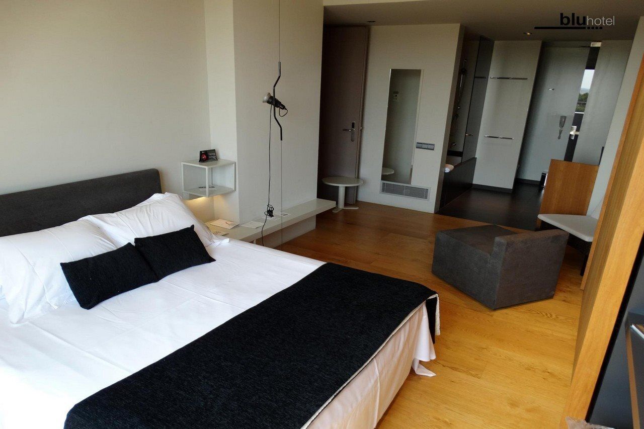 Habitación de hotel moderna con una cama grande, una mesita de noche, una lámpara, un espejo, piso de madera y una pequeña mesa redonda con una silla.