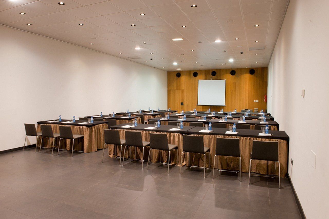 Un salón de conferencias con varias mesas y sillas, equipado con botellas de agua y documentos, y una pantalla de proyector en la pared.