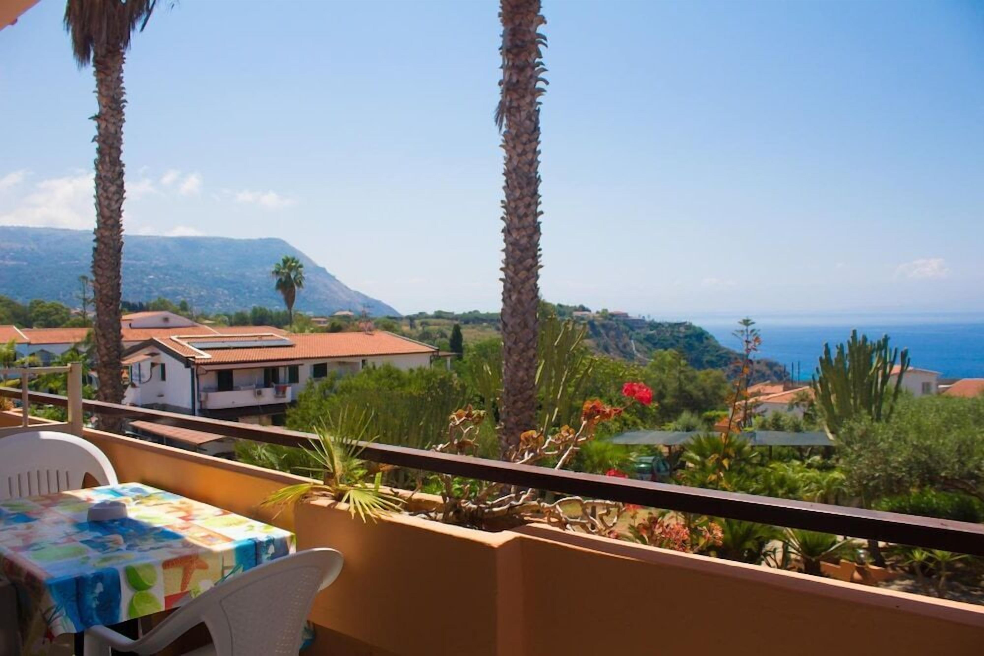 A balcony with a table and chairs, overlooking a coastal landscape with houses and palm trees.