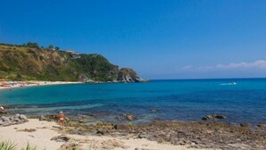 Una spiaggia con acque turchesi cristalline, costa rocciosa e un edificio in lontananza su una collina.