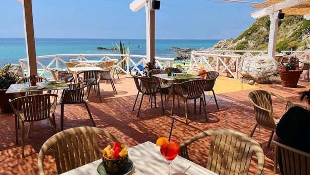 Un ristorante sul mare con posti a sedere all'aperto, vista sull'oceano e un tavolo con un cocktail tropicale e frutta fresca.