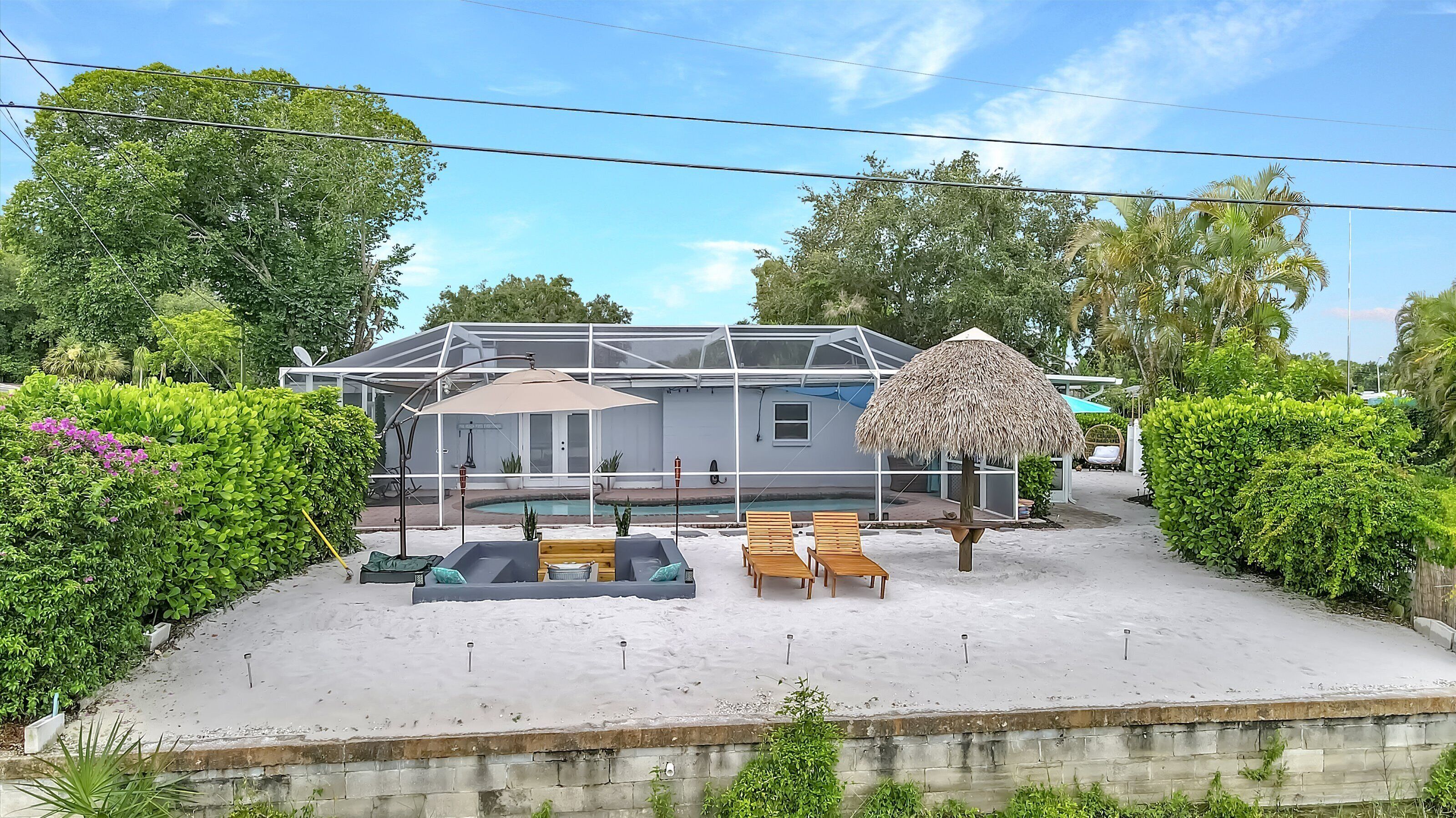 Una casa con piscina, rodeada de vegetación y una zona arenosa con sillas de descanso.