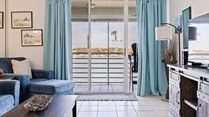 Una habitación con sofá, televisión, balcón con cortinas y vista a la playa.
