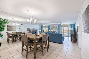 Ein modernes Wohnzimmer mit einem blauen Sofa, einem Essbereich mit einem Holztisch und Stühlen sowie einem großen Fenster mit Vorhängen.