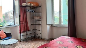 Een slaapkamer met een bed, een stoel, een klein tafeltje, een metalen plank en een raam met gordijnen.