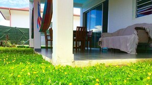 Ein überdachter Sitzbereich mit Patio-Tisch und -Stühlen, einer überdachten Couch und einem  üppigen, grünen Rasen mit gelben Blumen.