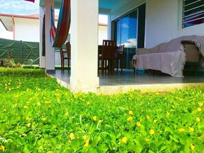 Ein überdachter Sitzbereich mit Patio-Tisch und -Stühlen, einer überdachten Couch und einem üppigen, grünen Rasen mit gelben Blumen.