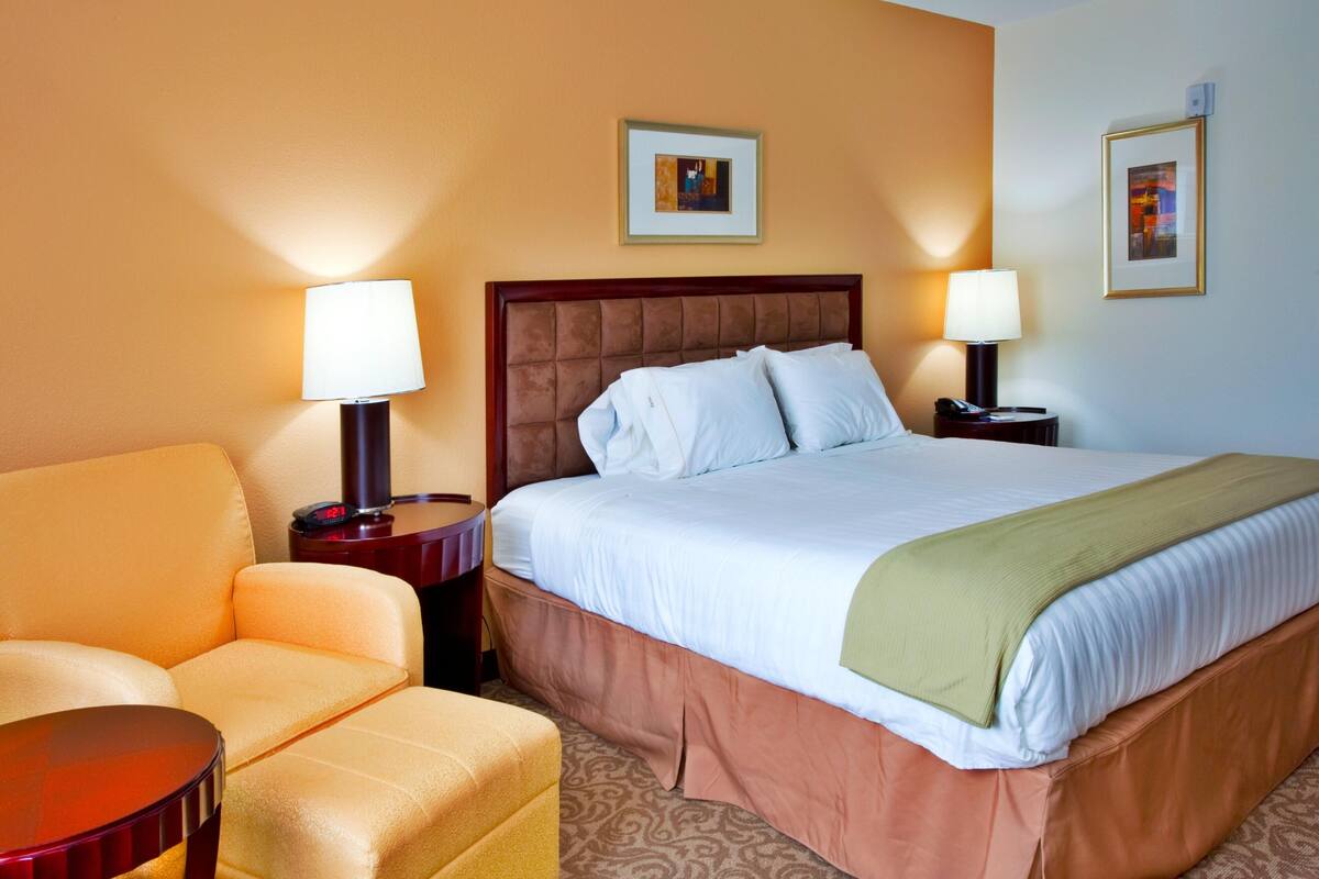 Une chambre d’hôtel avec un grand lit, deux tables de chevet avec des lampes, une chaise et deux tableaux encadrés au mur.