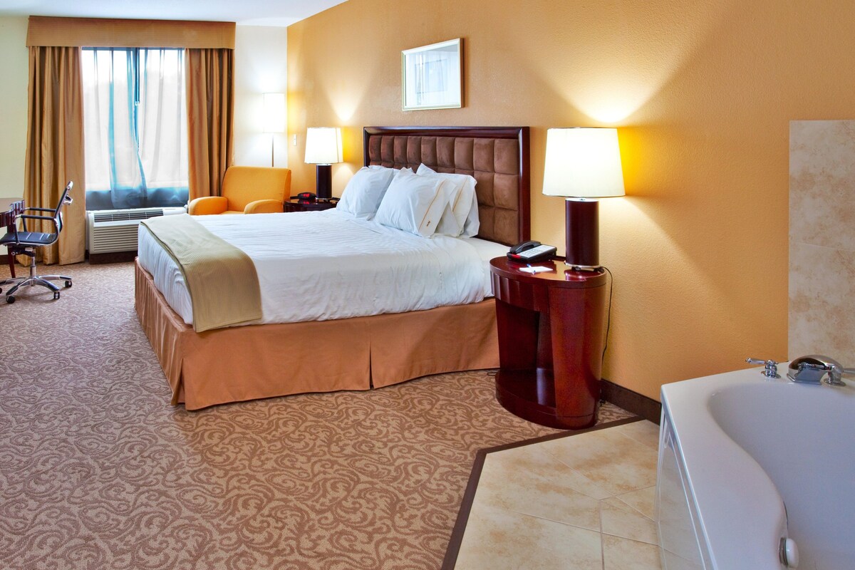 A hotel room with a large bed, two bedside lamps, a desk with a chair, a desk lamp, a telephone, and a bathtub.
