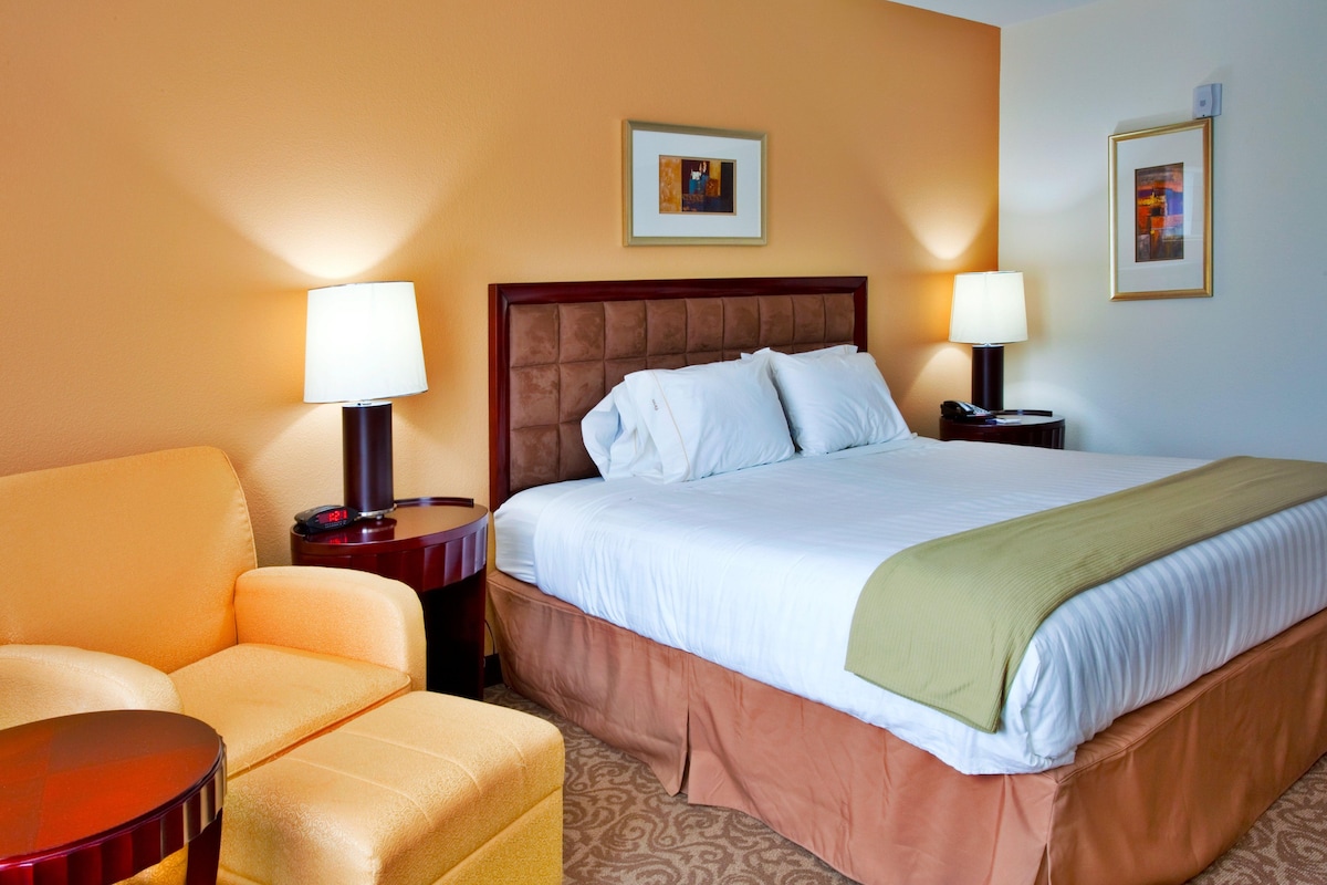 Habitación de hotel con una cama grande, dos mesitas de noche con lámparas, una silla y dos cuadros en la pared.