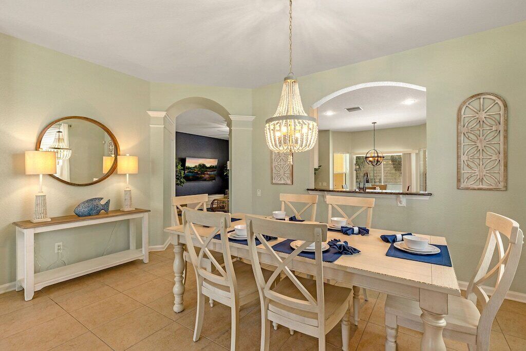 A dining area with a round table, white chairs, and a chandelier. There is a console table with lamps and a decorative fish.