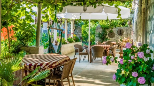 Un patio al aire libre con mesas y sillas, cubierto por una pérgola con plantas colgantes.