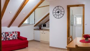 Un espacio habitable pequeño con un sofá rojo, una mesa de comedor de madera con un jarrón de flores, una zona de cocina con microondas y un espejo grande que refleja la habitación.