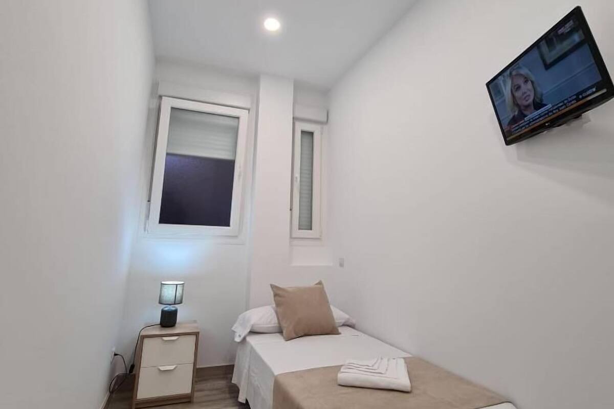 A small, well-lit bedroom with a single bed, a nightstand, a TV mounted on the wall, and a window with blinds.