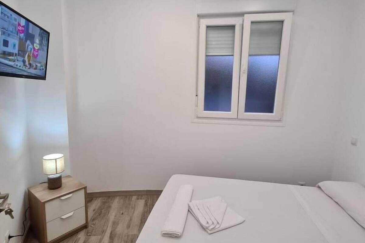 A hotel room with a bed, a nightstand with a lamp, a window with blinds, and a TV mounted on the wall.