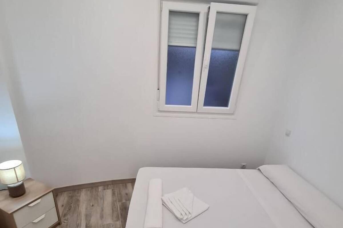 A small, clean room with a single bed, a nightstand, a lamp, and a window with blinds.