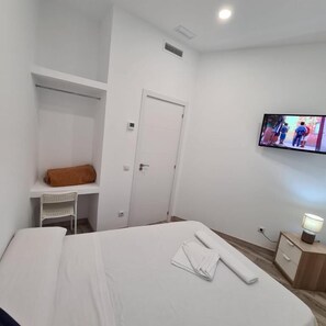 A hotel room with a bed, a TV mounted on the wall, a desk with a chair, and a wooden nightstand with a lamp. - Hostal Las Fronteras (Torrejón de Ardoz)