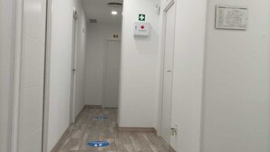 A clean, well-lit hallway with a wooden floor, white walls, and doors on both sides. There are blue directional markers on the floor, and a first aid kit is mounted on the wall. - Hostal Las Fronteras (Torrejón de Ardoz)