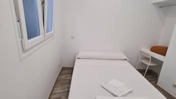 Quarto pequeno e limpo, com uma cama de solteiro, janela com persianas, escrivaninha e cadeira.