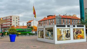 Una plaza pública con un edificio moderno, una bandera y un quiosco con obras de arte.
