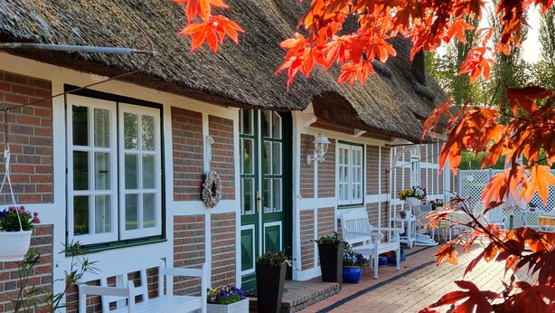 En traditionel bygning med stråtækt tag, hvide rammer om vinduer og døre, facade af mursten og udendørs siddepladser med plantekasser.