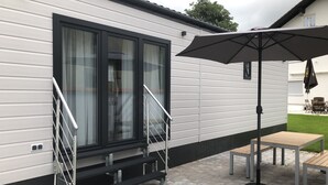 Een moderne stacaravan met een overdekte patio, compleet met een tafel, stoelen en een grote parasol, met een trap die naar een schuifdeur leidt.