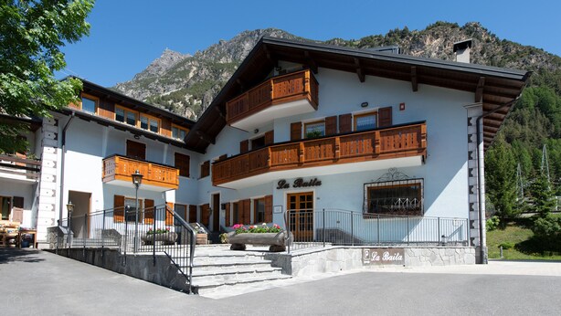 Un bâtiment à deux étages doté d’un balcon, d’une enseigne indiquant « La Baita », et ayant une chaîne de montagnes en toile de fond.