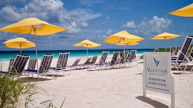 Une plage bordée de rangées de chaises longues et de parasols jaunes, un panneau indiquant «Valentines Resort» et affichant un message à l’attention des clients.