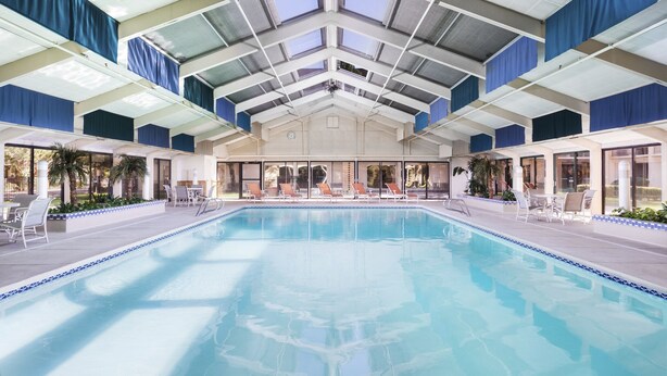 Une piscine intérieure à l’eau d’un bleu cristallin, entourée de chaises longues et surmontée d’un toit en verre.