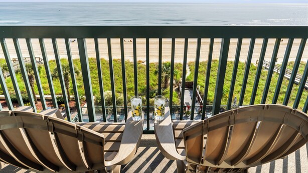 A balcony with two chairs and two glasses of water, overlooking a beach with people and greenery.