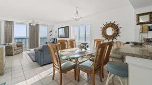 A spacious living area with a glass-top dining table surrounded by wooden chairs, a sofa, and a view of the ocean. - Ashworth 1101 - Ocean Front Condo - 4 Bed/3 Bath (North Myrtle Beach)