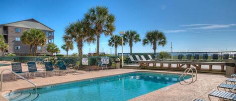 A swimming pool with lounge chairs and palm trees, surrounded by a fence and a building in the background.