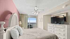 A bedroom with a bed, a ceiling fan, a dresser, a television, and a balcony with a view.