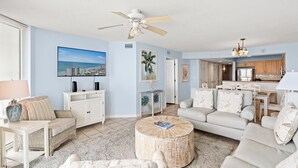 A spacious living room with a large flat-screen TV, a ceiling fan, and a kitchen area visible through an open doorway.