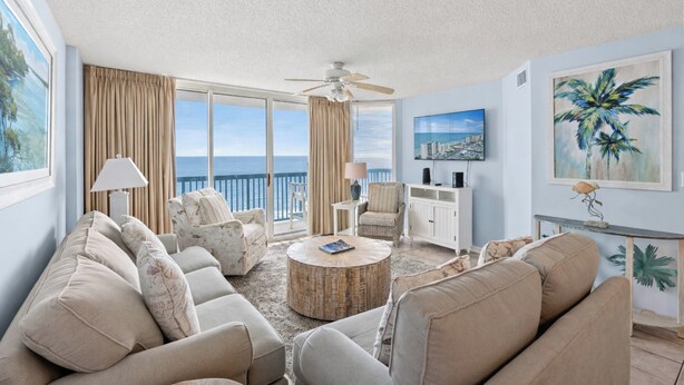 A spacious living room with a large window overlooking the sea, a ceiling fan, and a round coffee table.
