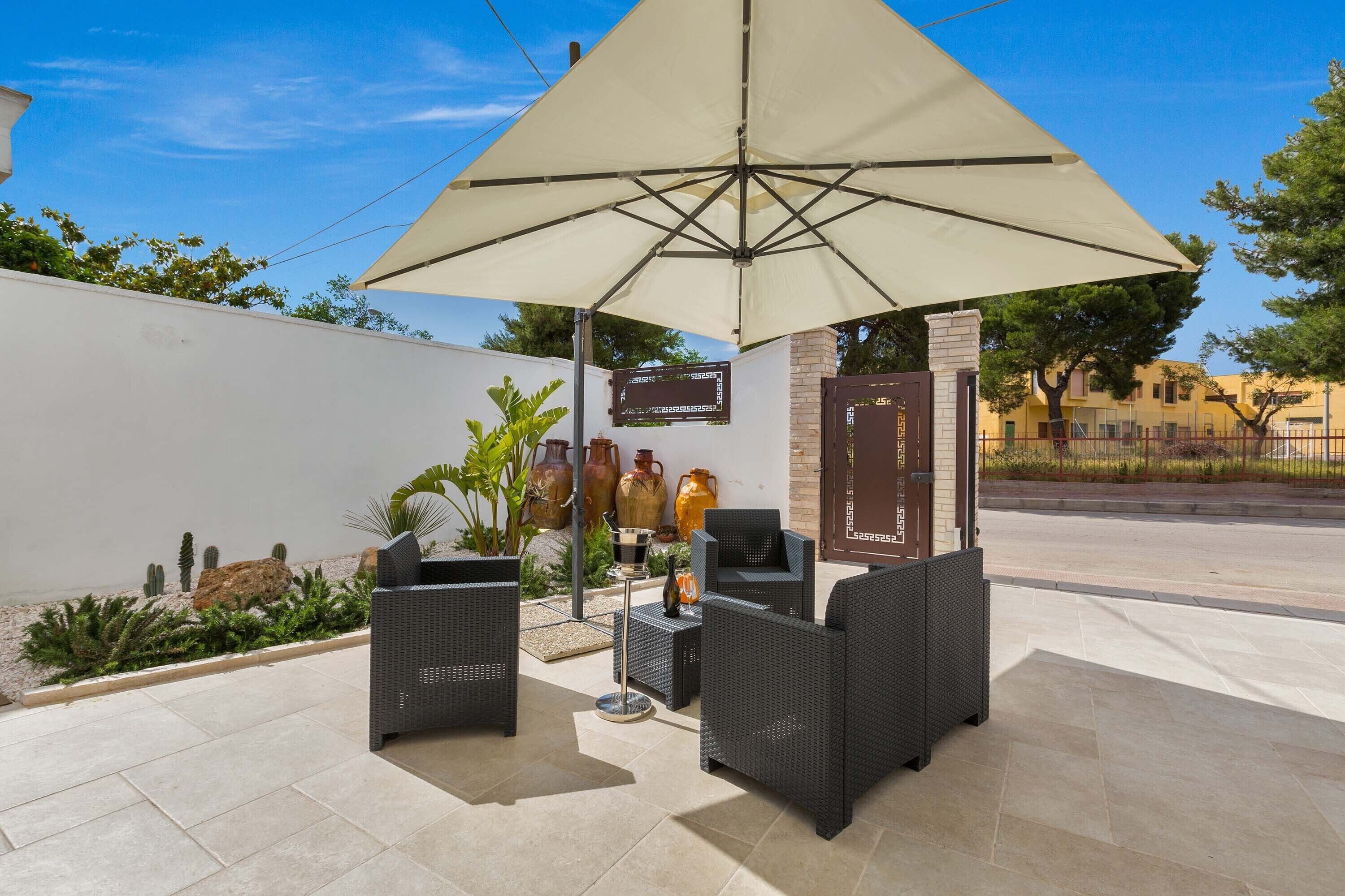 Un patio al aire libre con mobiliario de mimbre, un paraguas grande y plantas decorativas.