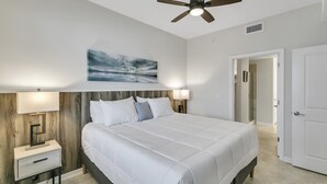 Une chambre à coucher comprenant un lit, des lampes de chevet, un ventilateur de plafond, un tableau et une porte donnant sur une salle de bain.