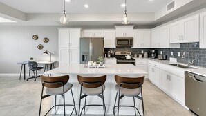 Une cuisine moderne avec des meubles blancs, un îlot central doté de tabourets de bar, des appareils en acier inoxydable et un éclairage suspendu.