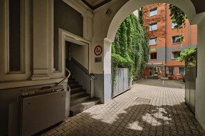 Un pasillo con una escalera que conduce a un piso superior, un cartel que indica un área de estacionamiento y vistas de un edificio de fachada de ladrillo y vegetación.