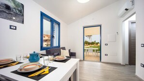 Ein moderner Essbereich mit einem weißen Tisch für zwei Personen, einem blauen Topf und Blick durch eine Glastür auf eine Terrassen-Sitzgruppe.