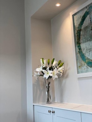 Un vase blanc orné de fleurs blanches, posé sur un meuble blanc dans une pièce aux murs blancs, où l’on aperçoit également un tableau.