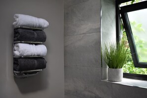 Une salle de bain avec un porte-serviettes où sont rangées des serviettes blanches et foncées, une plante en pot sur le rebord d’une fenêtre et une vue sur la verdure à l’extérieur.