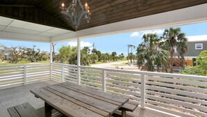 Eine überdachte Terrasse mit Holztisch und -bänken, weißen Geländern und Blick auf Palmen und eine Straße.