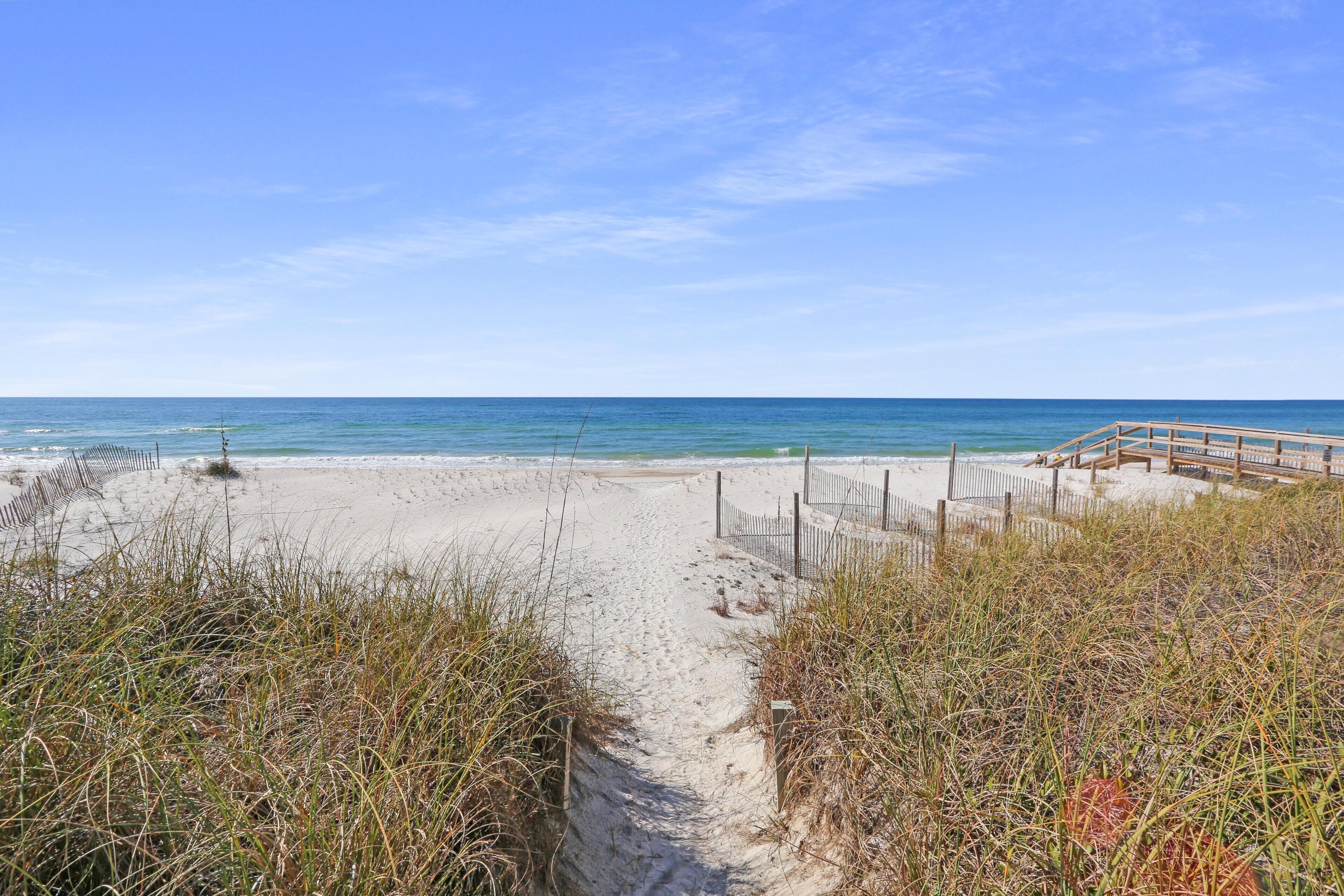 Un sentier de sable qui longe la plage et mène à une jetée en bois, elle-même bordée d’une clôture.