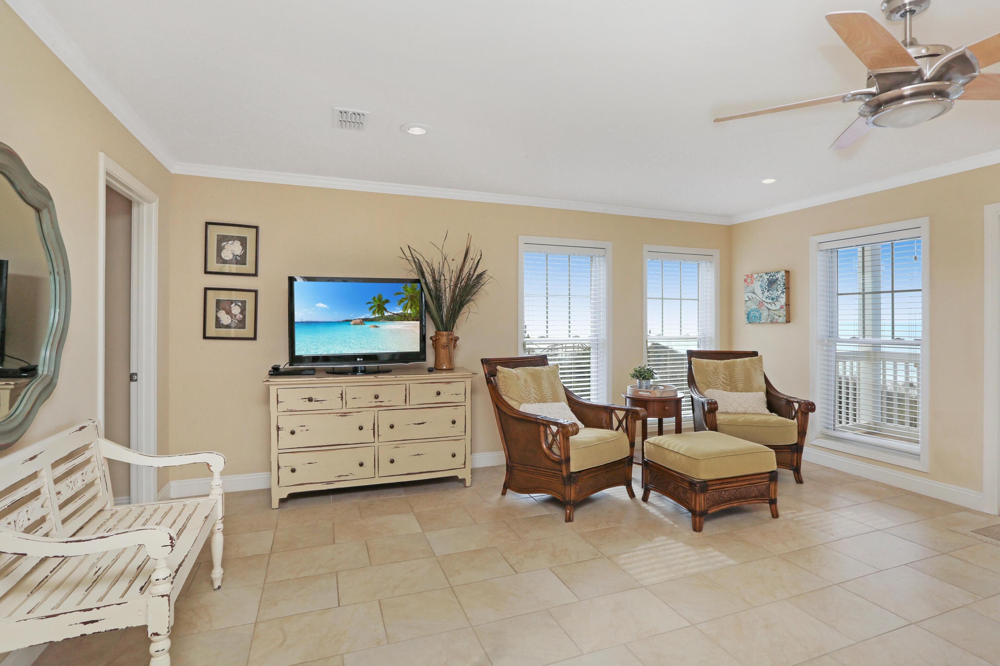 A living room with a television, a white bench, two armchairs, a wooden cabinet, and a ceiling fan.
