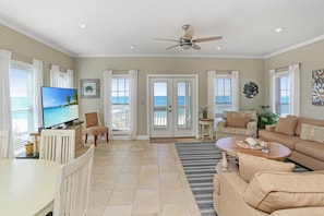 A spacious living room with a large flat-screen TV, comfortable seating, and a view of the ocean through the glass doors.