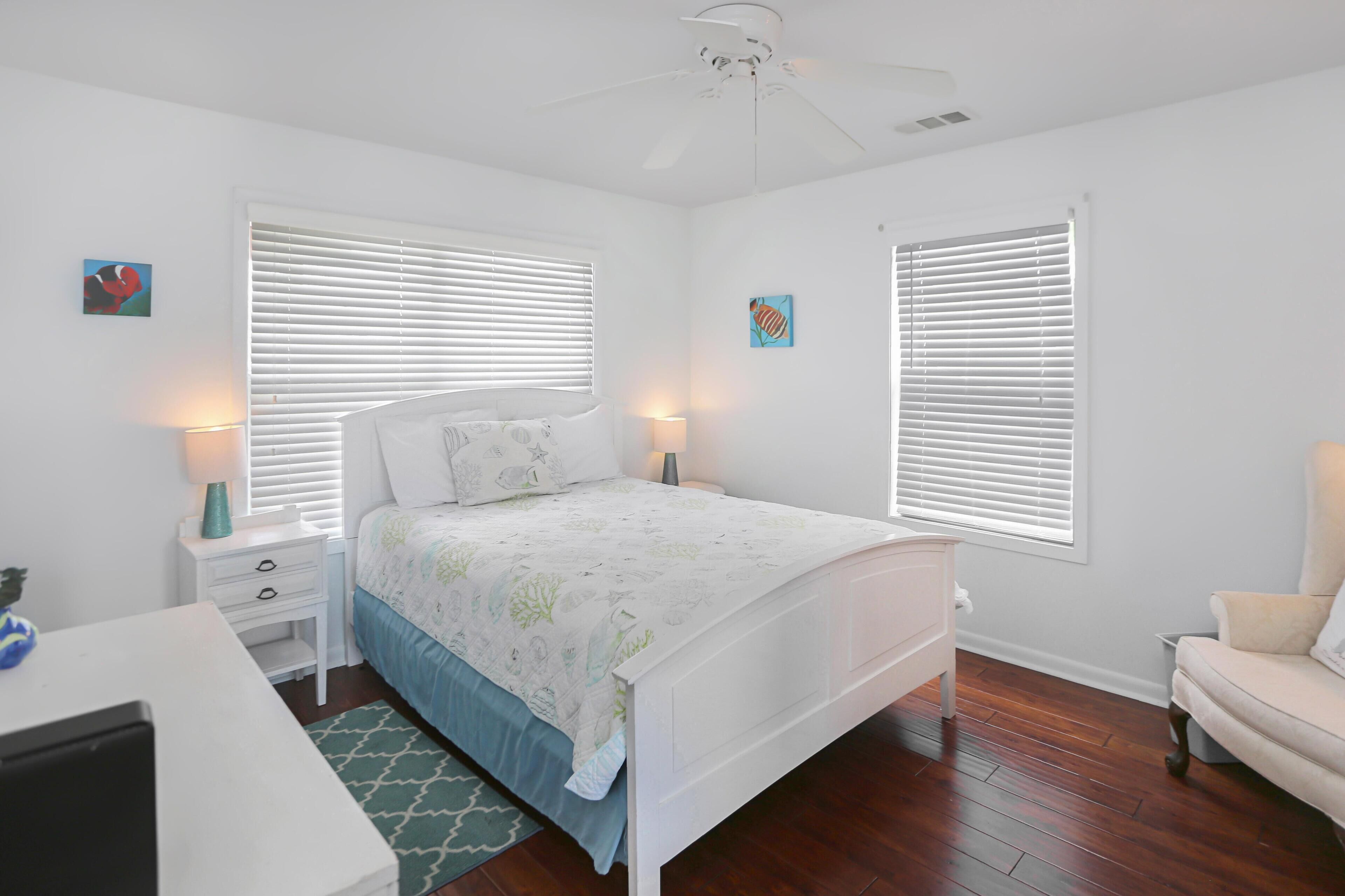 A bedroom with a bed, two bedside tables, a desk, a chair, and two windows with blinds.