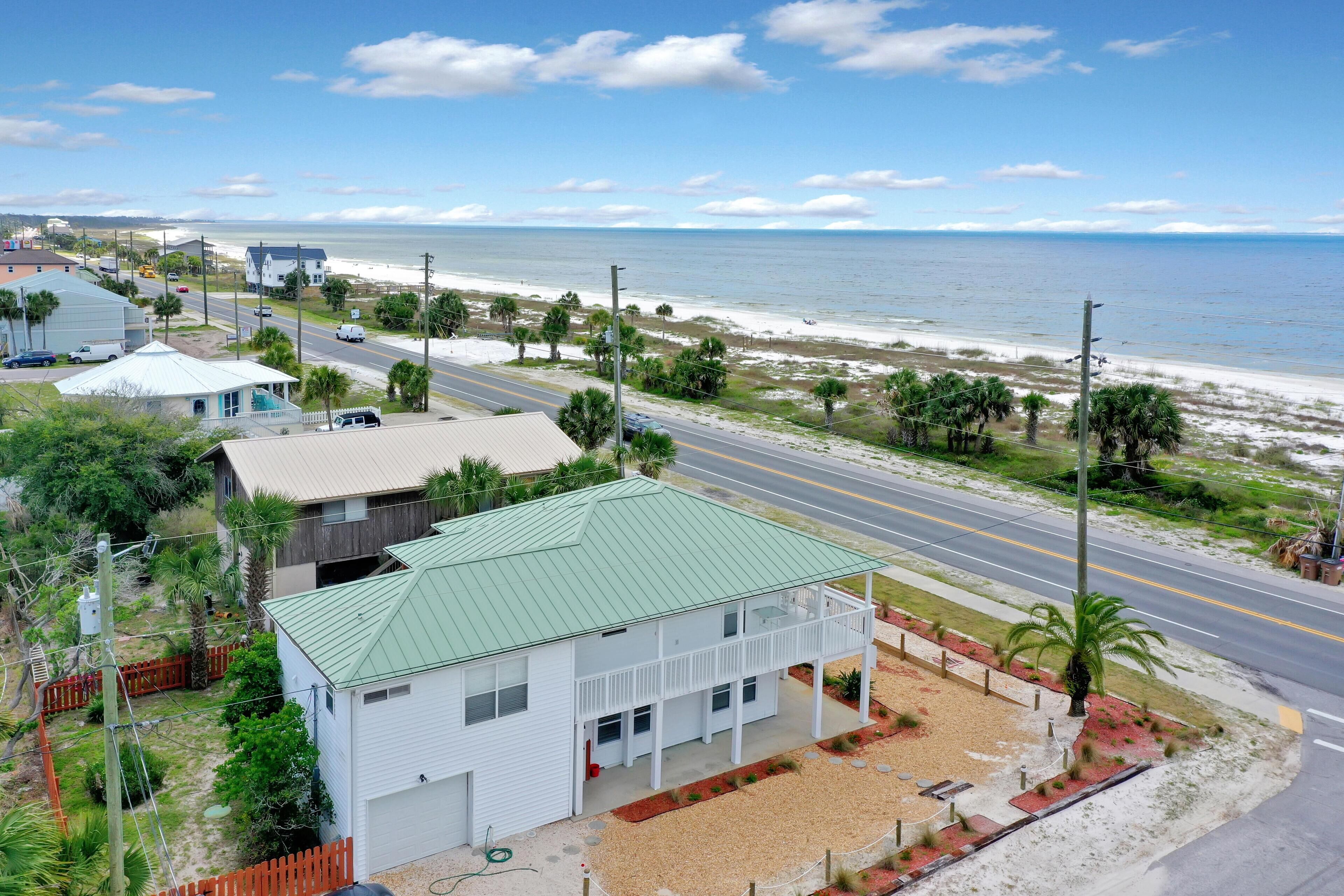 A coastal house with a green roof, a fenced yard, and a road leading to the beach.