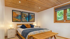 Une chambre avec un plafond en bois, un lit recouvert d’une couette à carreaux bleus et blancs, deux oreillers, une table de chevet avec une lampe et une fenêtre donnant sur un paysage verdoyant.