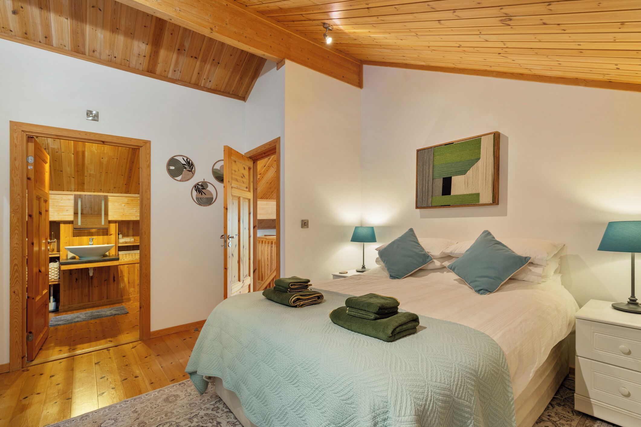A bedroom with a bed, two bedside lamps, a wooden wardrobe, and a bathroom with a sink and mirror.